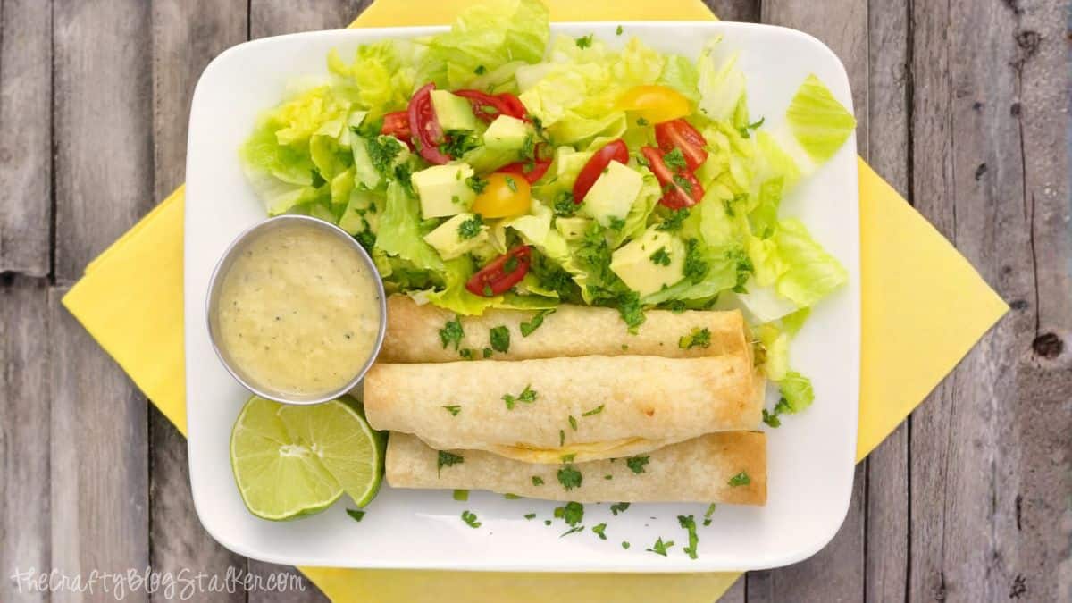 When I put Oven Baked Chicken Taquitos on the menu all my kiddos get so excited. I have been making these taquitos for a few years and they are always a crowd pleaser. Plate with oven-baked chicken taquitos, fresh lettuce salad topped with avocado and tomatoes, lime wedges, and creamy dipping sauce on the side.
