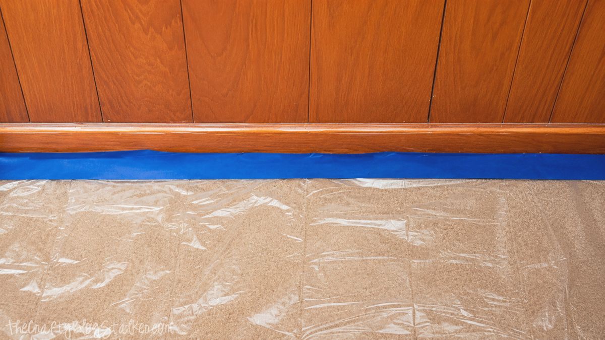 Plastic drop cloth taped along carpet edge with painter's tape to protect flooring before painting wood baseboards and paneling.