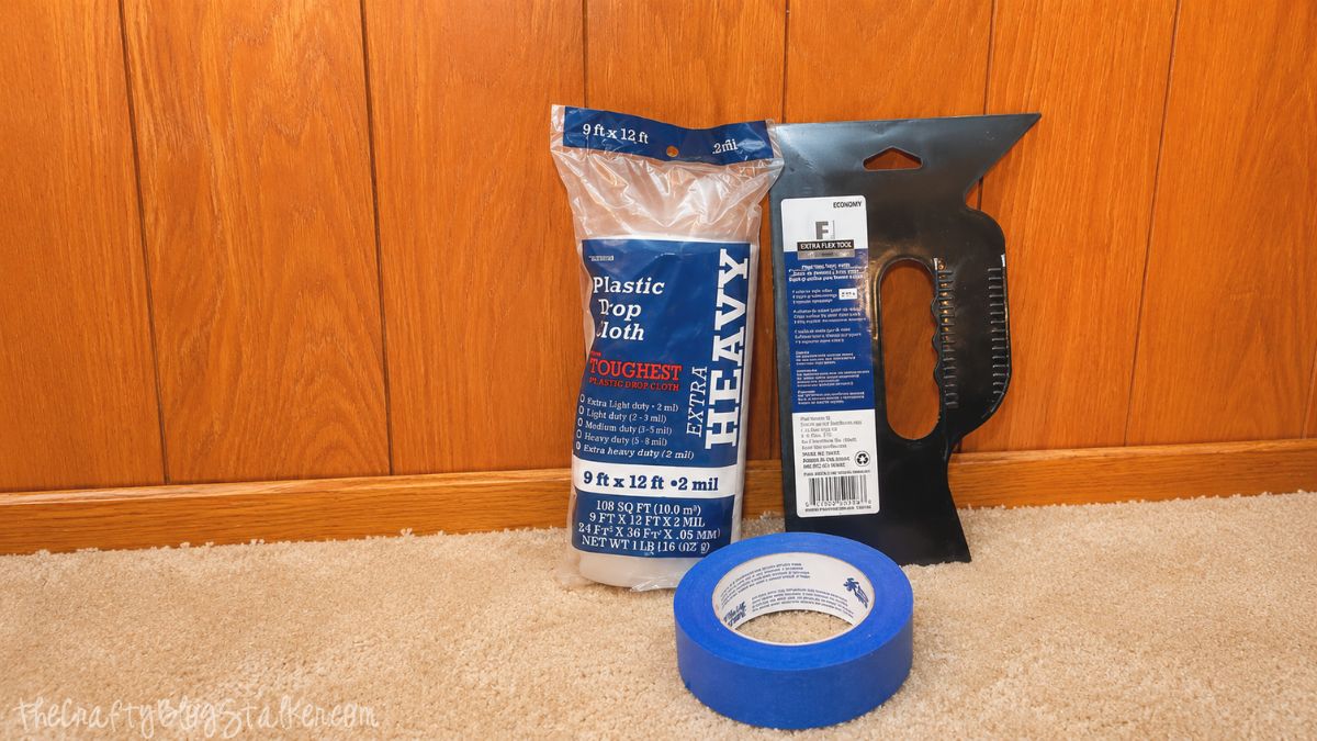 Painting supplies including a plastic drop cloth, painter's tape, and a paint edger tool arranged on carpet in front of wood paneling.