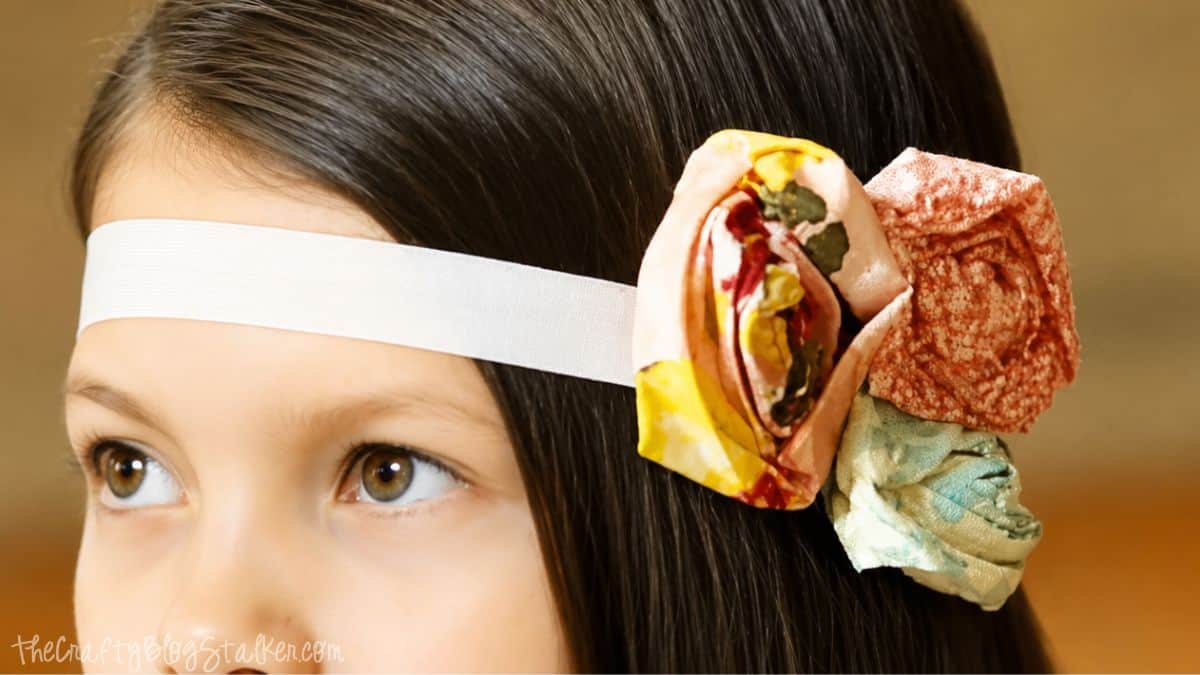 Young girl wearing a white elastic headband with colorful ruched fabric flowers on the side against a soft blurred background.