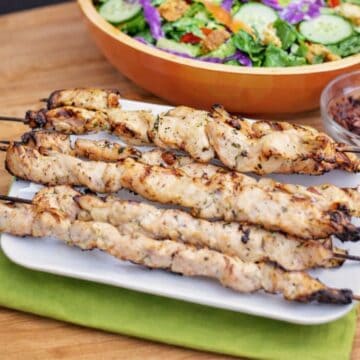 Grilled chicken skewers on a plate with a bowl of salad and dressing in the background on a wooden table.