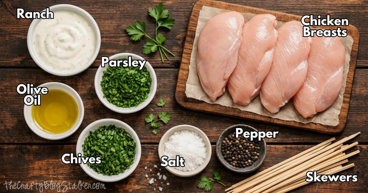 Ingredients for chicken skewers including chicken breasts, ranch dressing, olive oil, parsley, chives, salt, pepper, and wooden skewers on a table.