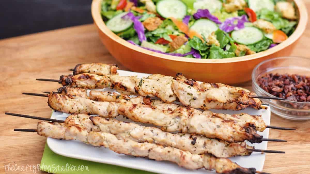 Grilled ranch chicken skewers on a white plate, served with a colorful garden salad and a bowl of bacon bits on a wooden table.