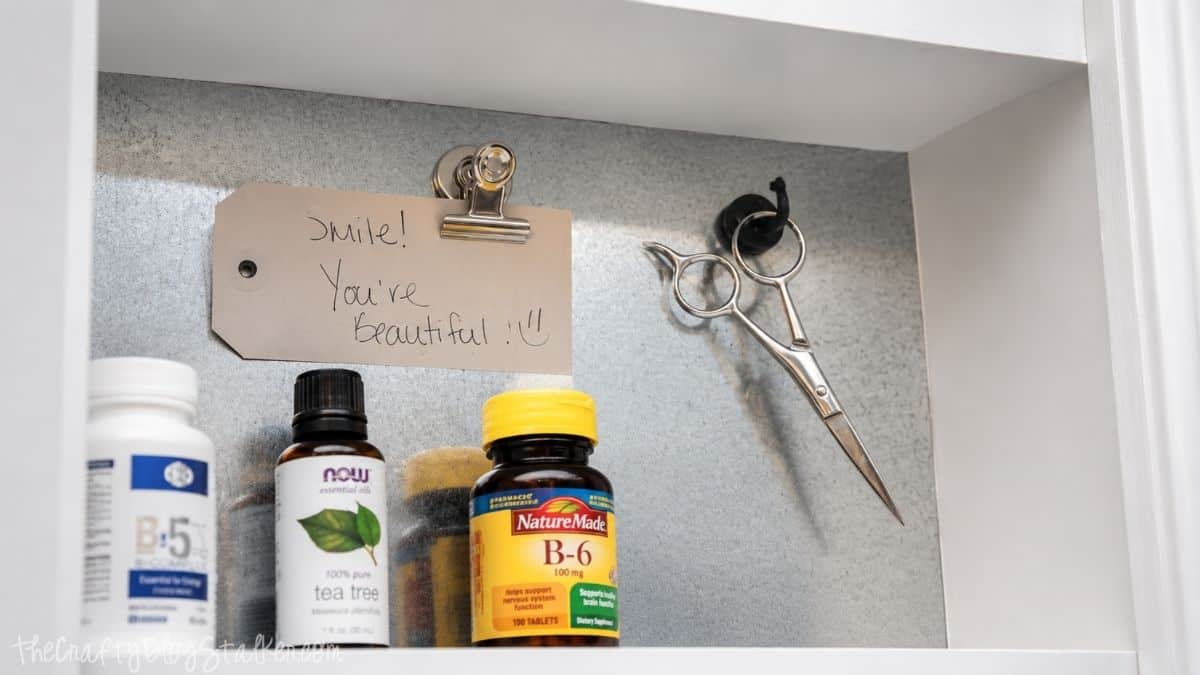 Magnetic medicine cabinet with a handwritten note, scissors on a magnetic hook, and organized vitamins and essential oils on the shelf.