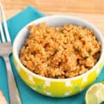 Bowl of restaurant-style Mexican rice garnished with cilantro beside a lime wedge on a blue napkin with a fork on a wood table.