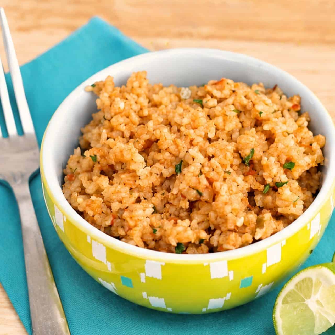 Bowl of restaurant-style Mexican rice garnished with cilantro beside a lime wedge on a blue napkin with a fork on a wood table.
