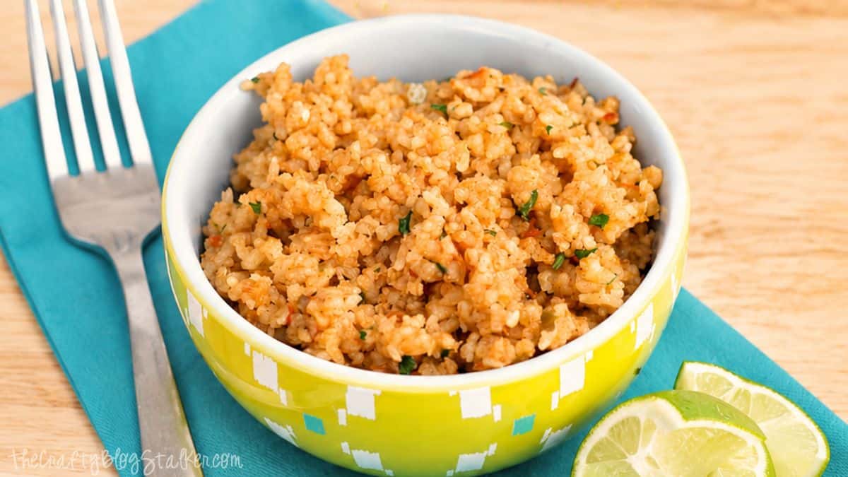 I have tried many recipes, but this recipe is hands down the best Mexican rice I have ever had. Bowl of Mexican rice on a teal napkin with a fork and lime wedges on a wooden table.