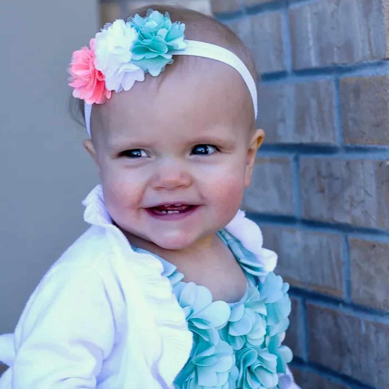Smiling baby wearing a colorful flower headband, white cardigan, and turquoise floral outfit while sitting near a brick wall.