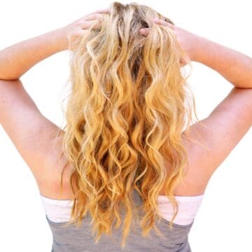 Back view of a woman with long wavy hair, hands lifting hair at the scalp to show texture and waves.
