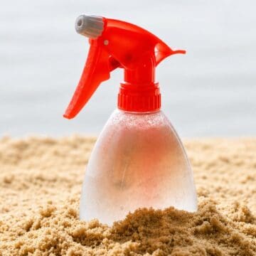 Clear spray bottle with a red nozzle sitting in sand, covered in condensation droplets.