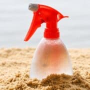 Clear spray bottle with a red nozzle partially buried in sand, covered in condensation droplets.