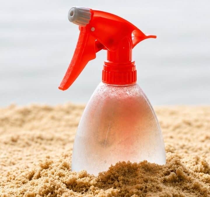 Clear spray bottle with a red nozzle partially buried in sand, covered in condensation droplets.