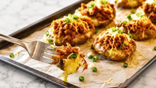 Fork holding a bite of BBQ chicken smashed potato topped with cheese and green onions, with more loaded smashed potatoes on a baking sheet in the background.