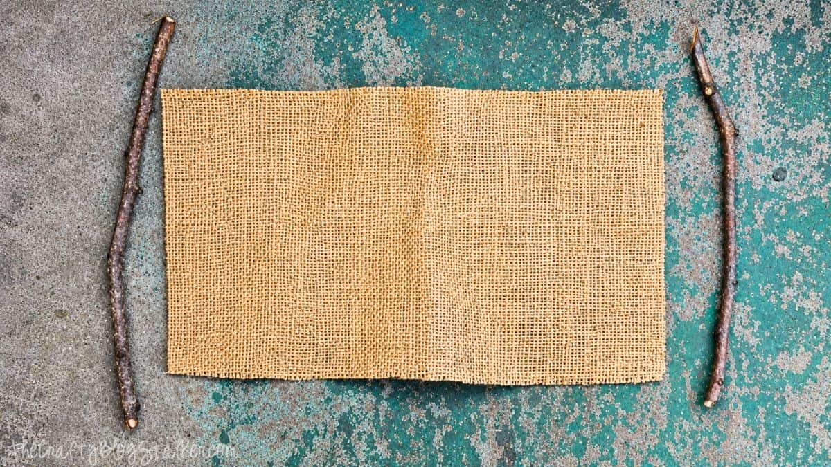Rectangle piece of burlap fabric with two small tree branches laid beside it on a weathered painted surface.