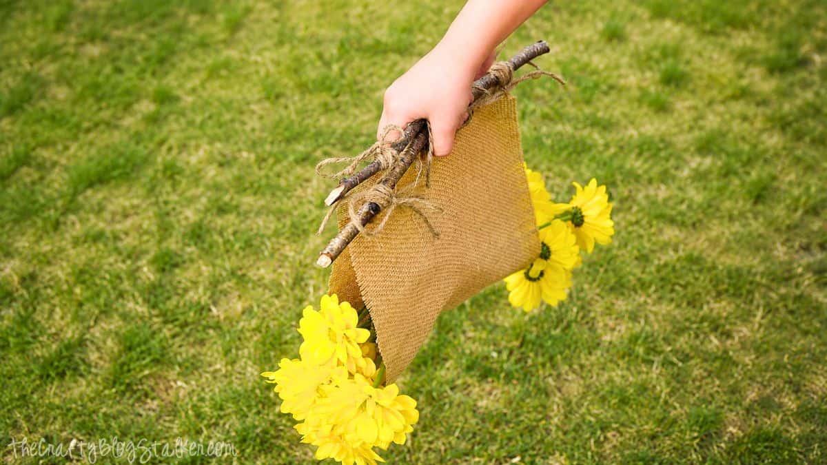 Hand holding a rustic burlap flower holder decorated with yellow flowers and tied branches while walking across a grassy yard.