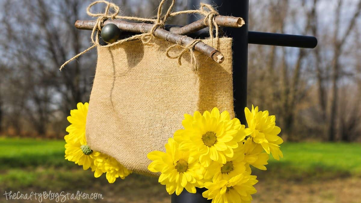Rustic burlap flower holder hanging outdoors with twine, branches, and bright yellow flowers attached to the sides.