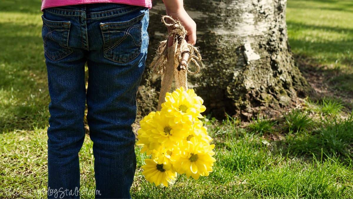 Child holding a rustic burlap flower holder filled with bright yellow flowers while standing near a tree in a grassy yard.