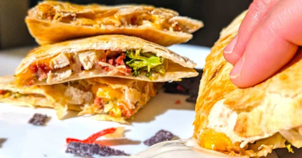 Stack of sliced quesadillas filled with chicken, bacon, cheese, and vegetables, with a hand holding one piece and dipping it into sauce.