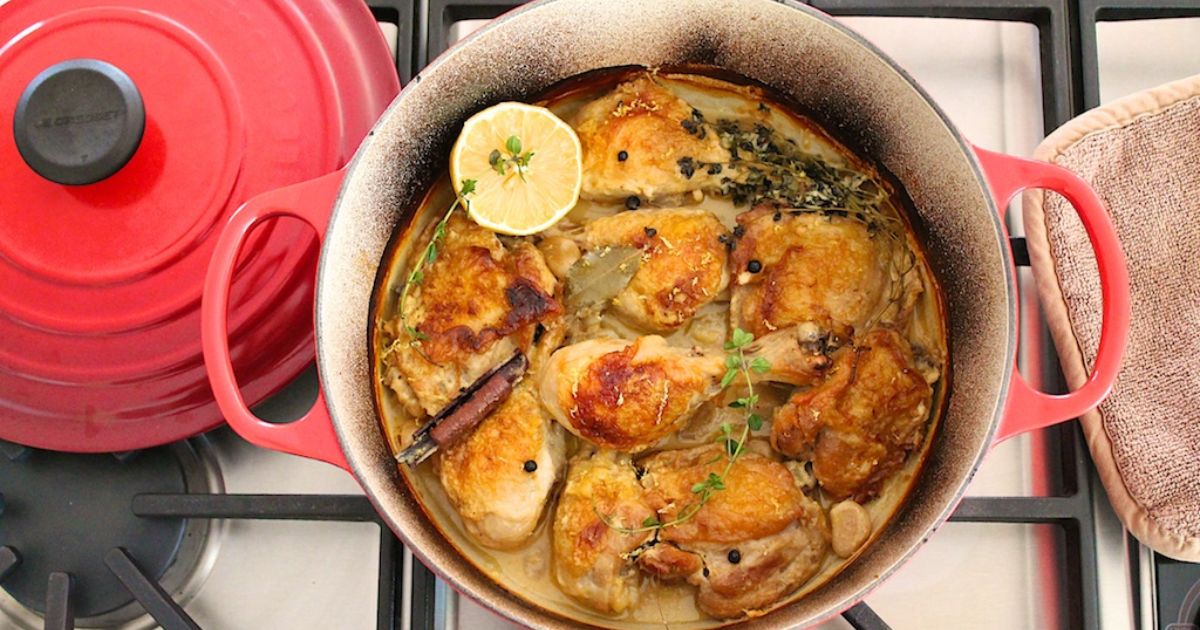 Dutch oven filled with browned chicken pieces simmering in broth with lemon, herbs, and garlic, set on a stovetop next to the lid.