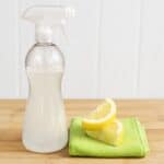 Spray bottle of homemade all-purpose cleaner beside lemon wedges and a green microfiber cloth on a wooden countertop.