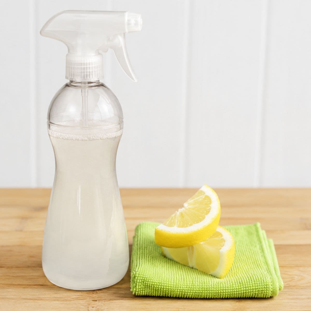 Spray bottle of homemade all-purpose cleaner beside lemon wedges and a green microfiber cloth on a wooden countertop.