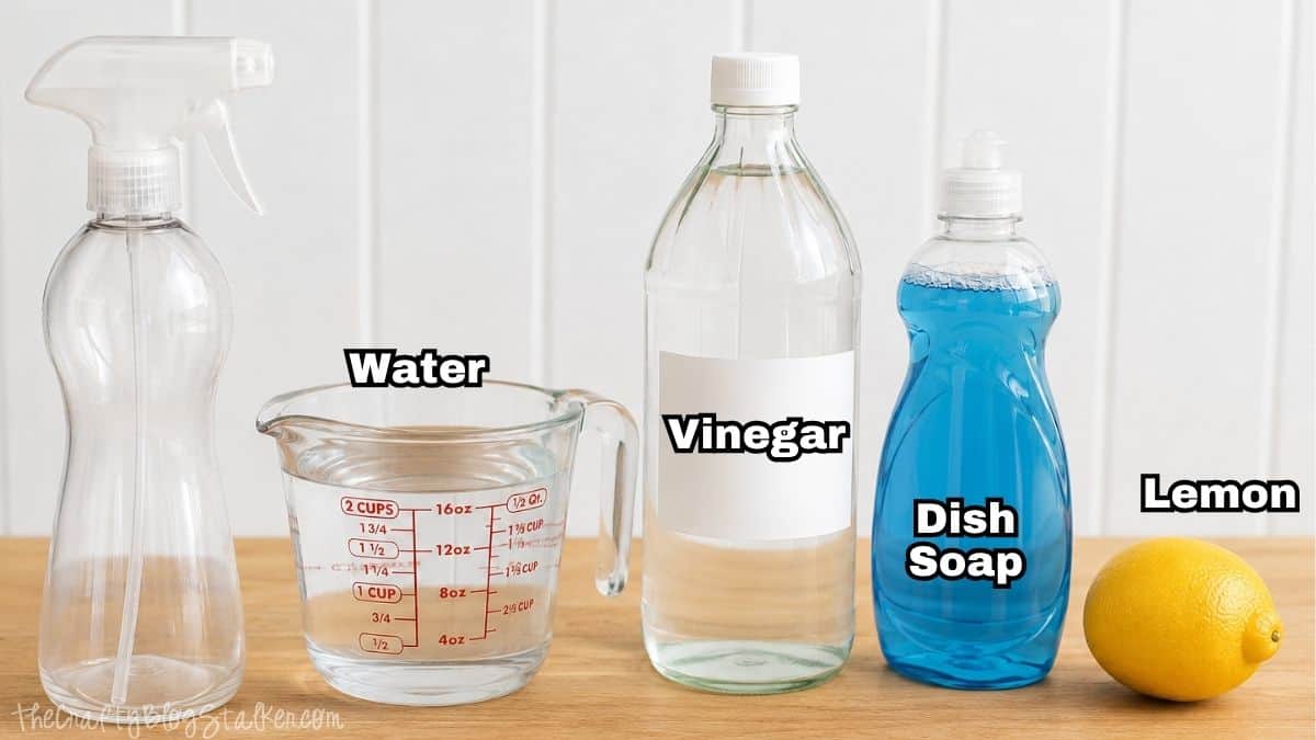 Ingredients for homemade all-purpose cleaner including an empty spray bottle, water, vinegar, dish soap, and a fresh lemon on a wooden surface.
