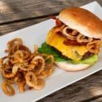 Double bacon cheeseburger topped with crispy onion rings and bacon, served with fried onion strings on a white plate outdoors.
