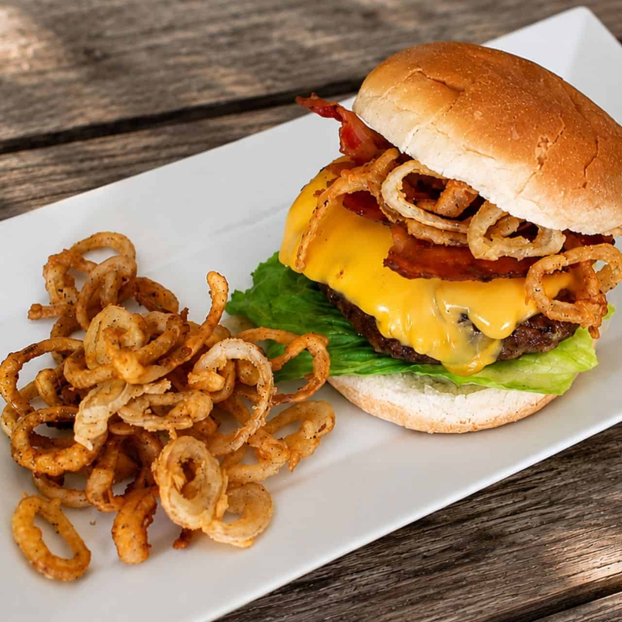 Double bacon cheeseburger topped with crispy onion rings and bacon, served with fried onion strings on a white plate outdoors.