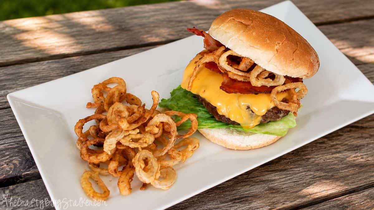 Double bacon cheeseburger with melted cheese, bacon, and crispy onion rings served with fried onion strings on a white plate outdoors.