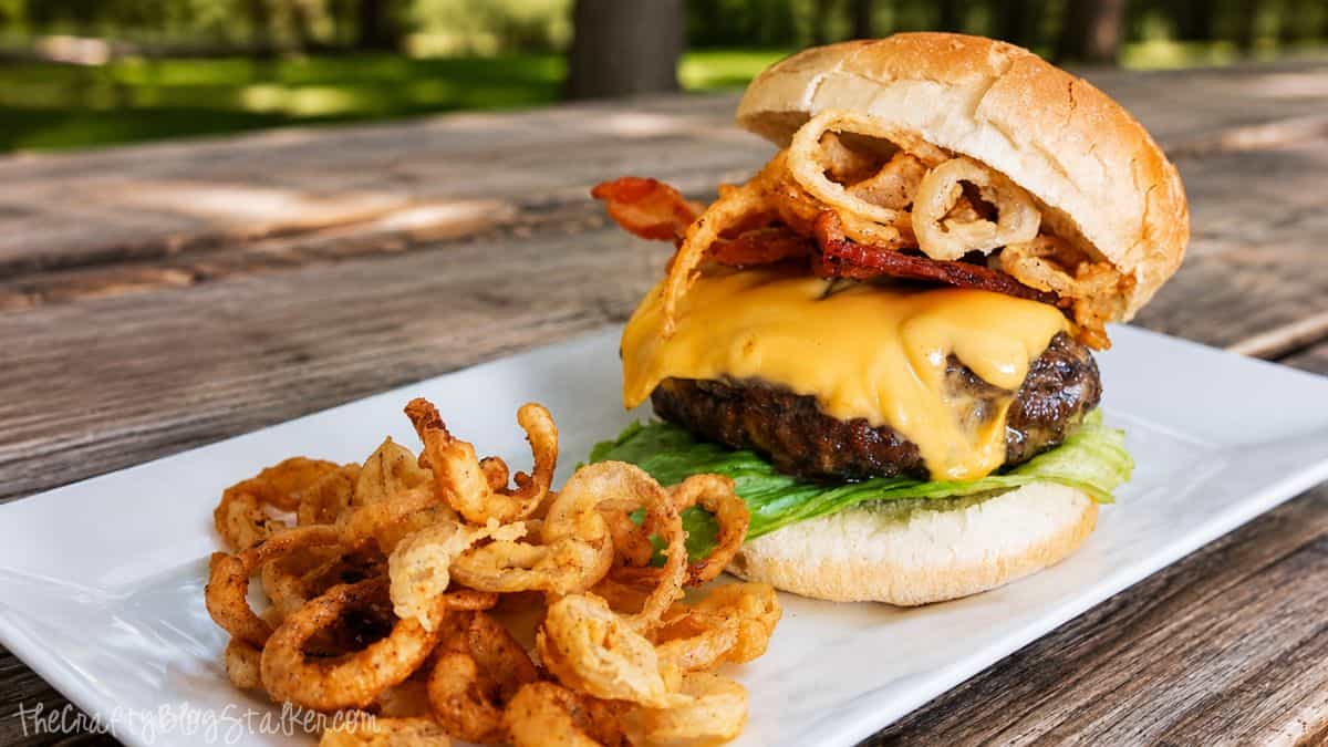 Double bacon cheeseburger topped with crispy onion rings and bacon, served with fried onion strings on a white plate outdoors.