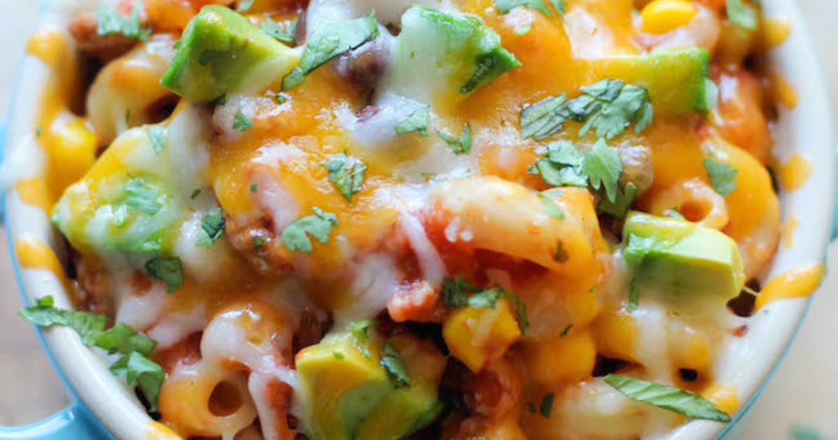 Close-up of pasta with melted cheese, avocado chunks, corn, and chopped herbs mixed together in a creamy, savory dish.