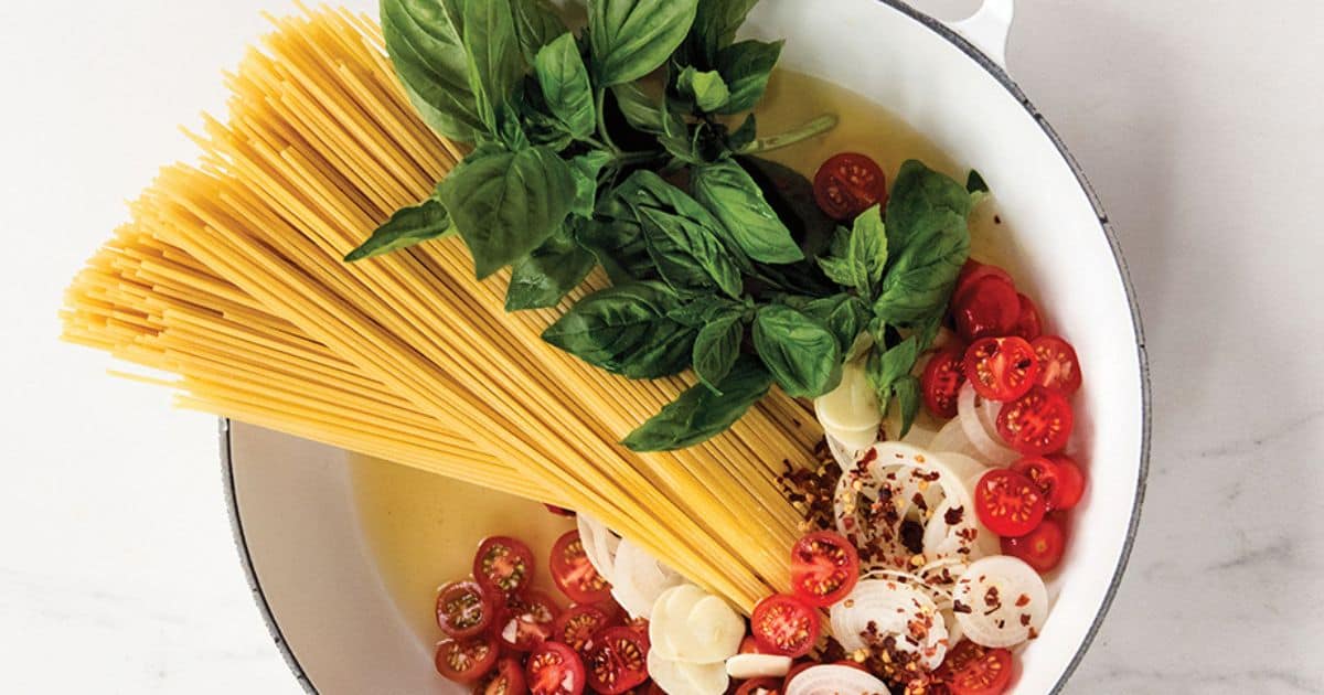 Uncooked spaghetti in a pot with cherry tomatoes, sliced onions, garlic, fresh basil, and seasonings arranged in broth before cooking.