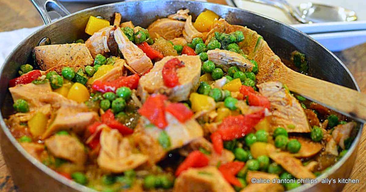 Skillet with sliced meat, peas, peppers, and diced vegetables mixed together in a savory sauce, with a wooden spoon stirring the dish.