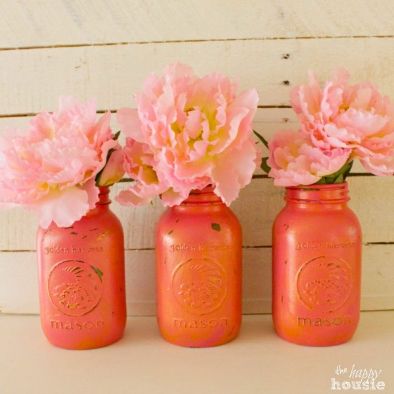 Three pink and gold painted mason jars filled with soft pink peony flowers displayed on a rustic white wood background.