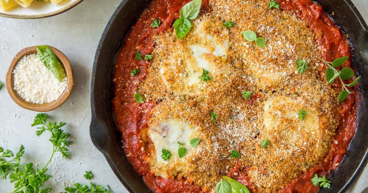 Skillet with breaded chicken topped with melted cheese in tomato sauce, garnished with fresh herbs and served alongside grated cheese and parsley.