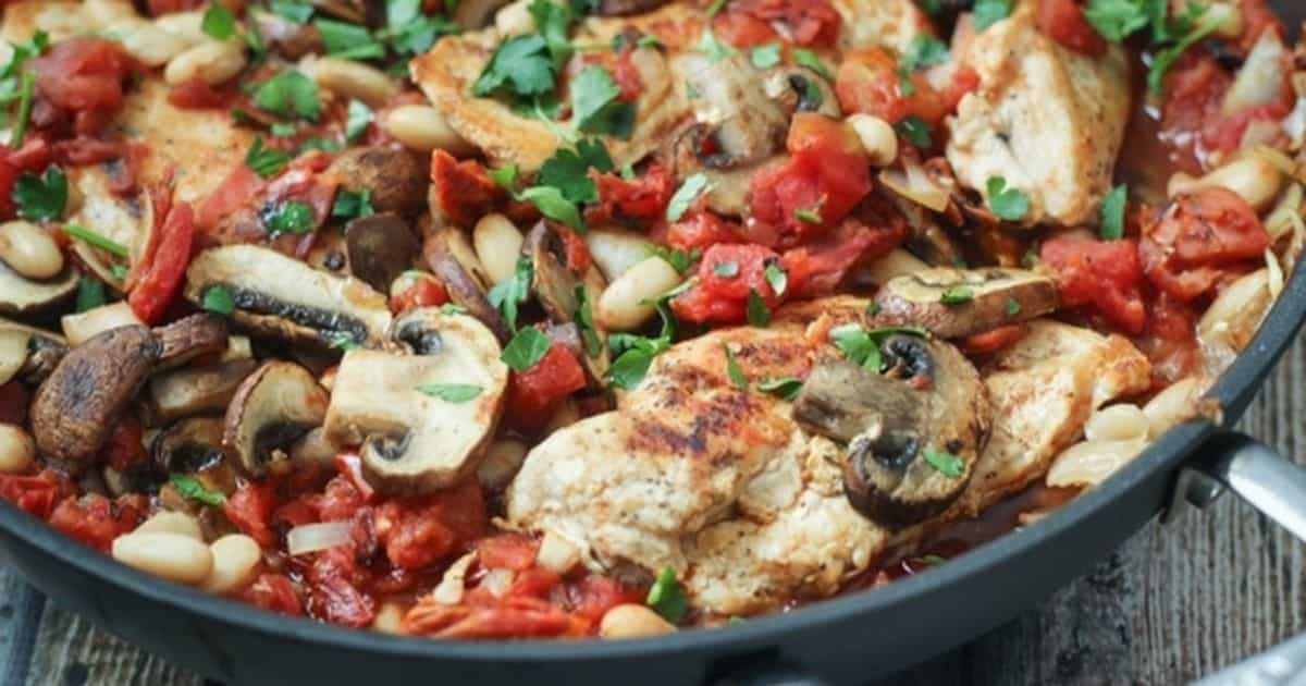 Skillet filled with sliced chicken, mushrooms, beans, and diced tomatoes topped with chopped herbs, cooking together in a savory sauce.