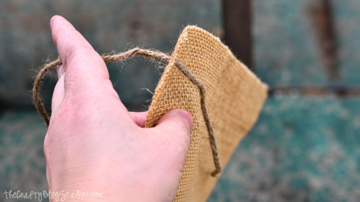 Hand holding burlap fabric while threading twine through the edge to create a rustic handmade flower holder.