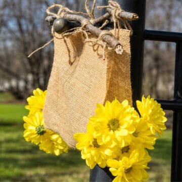 Rustic burlap flower holder tied to branches with twine and decorated with bright yellow flowers hanging outdoors on a black post.