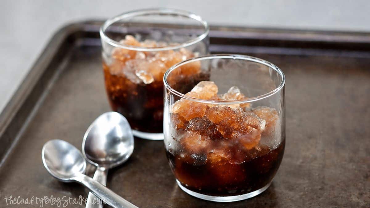 Two glasses of cola slush with ice crystals on a metal tray, with three spoons beside them.