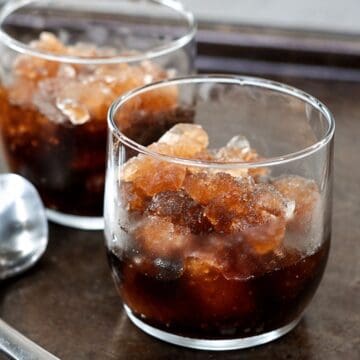 Two glasses of cola slush with ice crystals on a metal tray, with three spoons beside them.