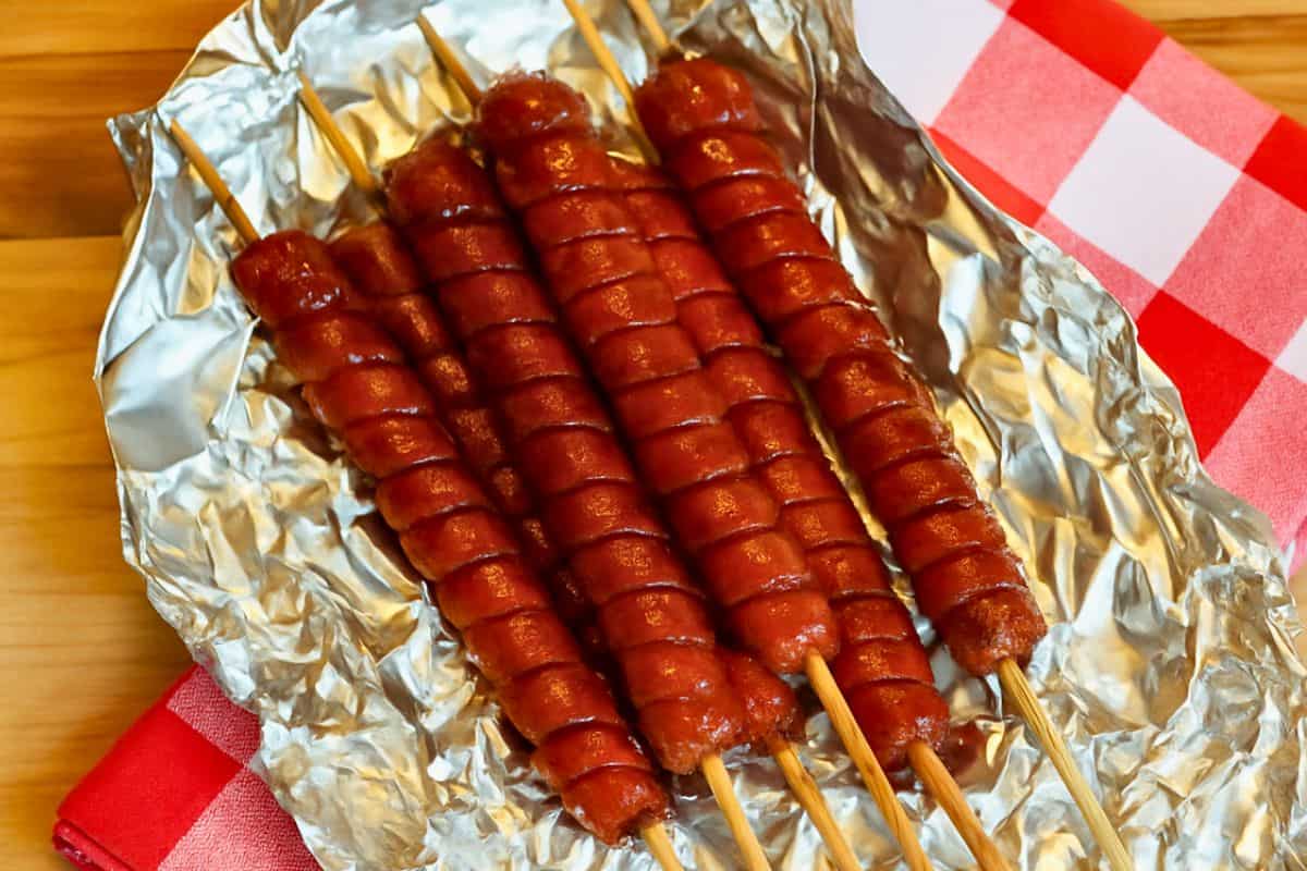 Five spiral-cut hot dogs on wooden skewers, grilled and served on foil over a red and white checkered napkin on a wooden surface.