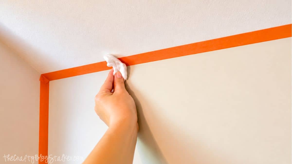 Hand pressing orange painter's tape along a slanted wall edge to prep a wall for chalkboard paint.