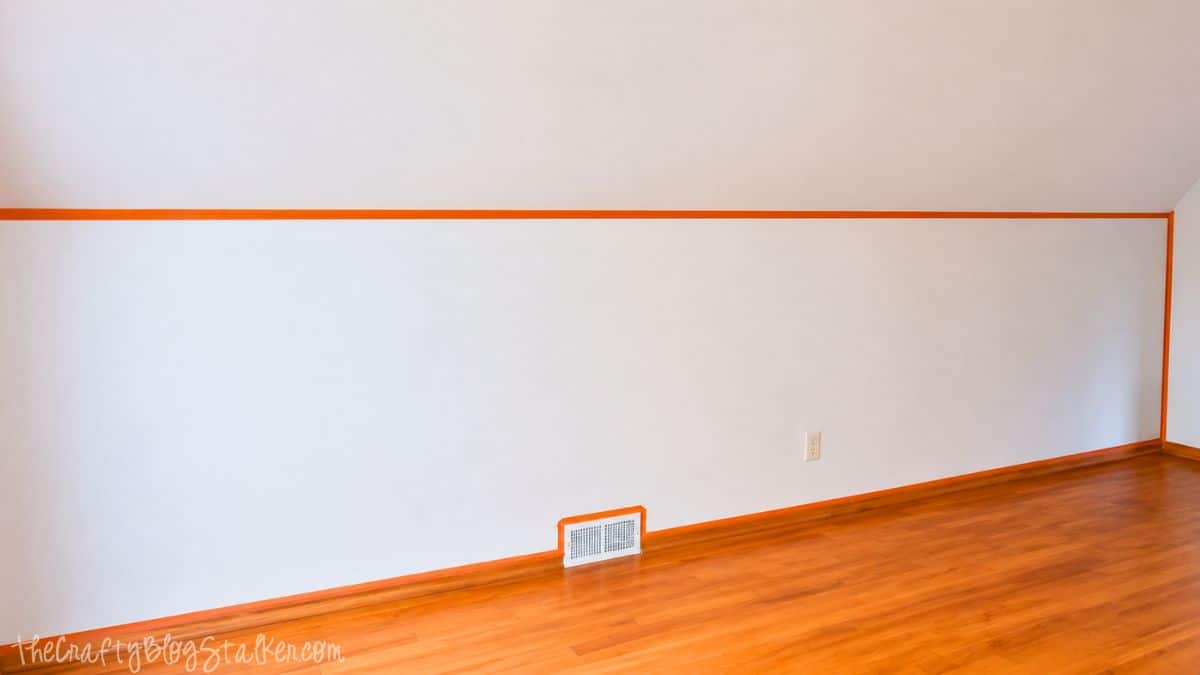 Wall taped off with orange painter's tape along the edges before applying chalkboard paint in a bedroom with hardwood floors.