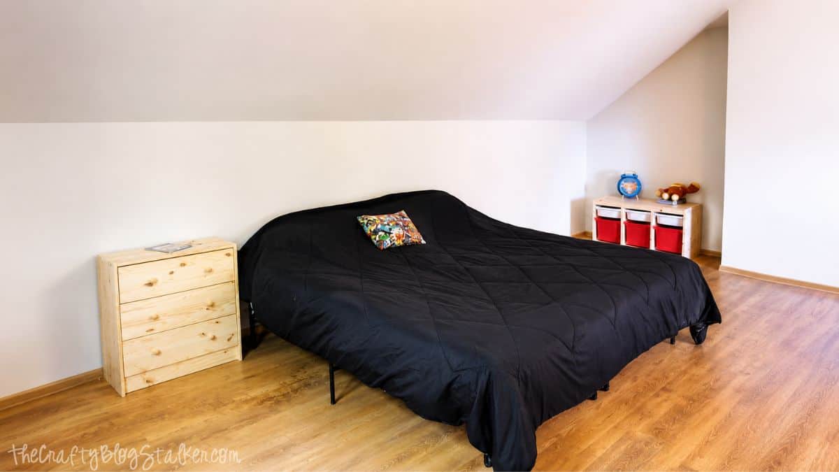 Large bedroom with slanted ceiling, black bedspread, wood floors, and minimal furniture before adding a chalkboard wall.