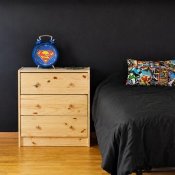 Bedroom corner with black chalkboard wall, wood nightstand, blue Superman clock, and bed with comic book pillow.