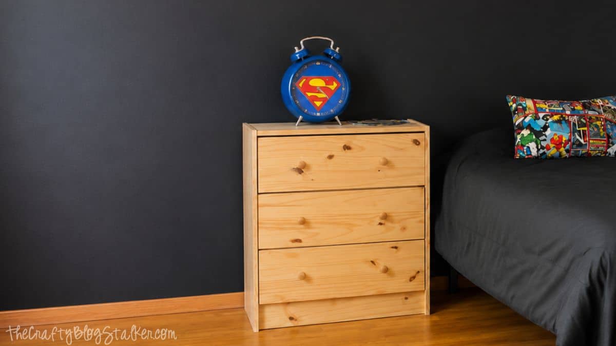 Wood nightstand beside a bed against a black chalkboard wall with a blue Superman clock and comic book accent pillow.