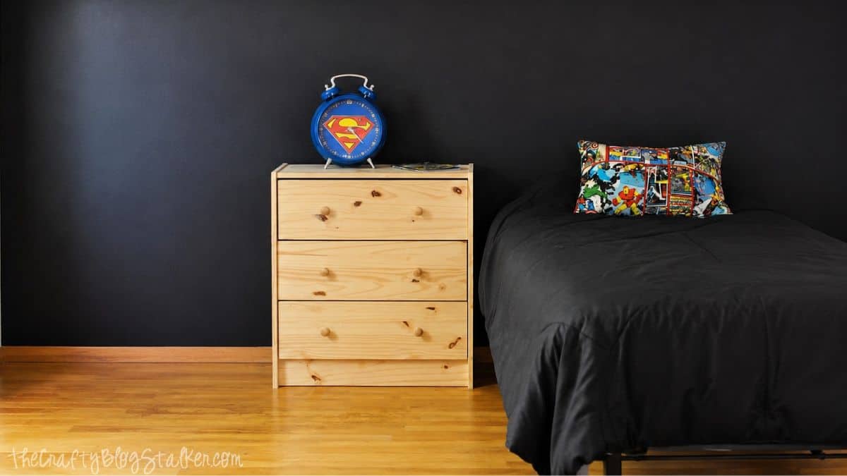 Bedroom with black chalkboard wall, wood nightstand, blue Superman clock, black bedding, and comic book accent pillow.