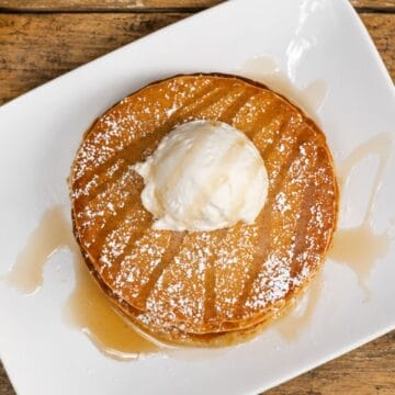 Stack of root beer pancakes topped with whipped cream, powdered sugar, and syrup on a white plate over a wooden table.