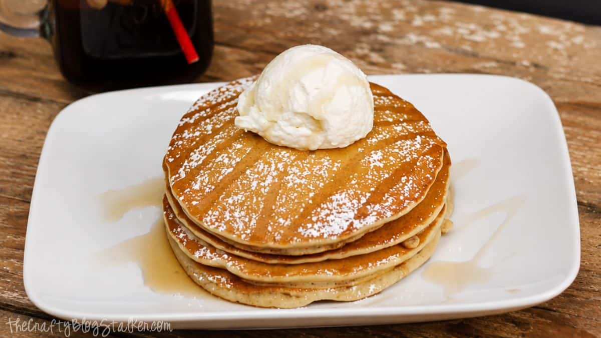 Stack of root beer pancakes topped with whipped cream, powdered sugar, and syrup on a white plate with a glass of root beer nearby.