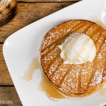 Stack of root beer pancakes topped with vanilla ice cream, powdered sugar, and syrup on a white plate beside an iced root beer drink.